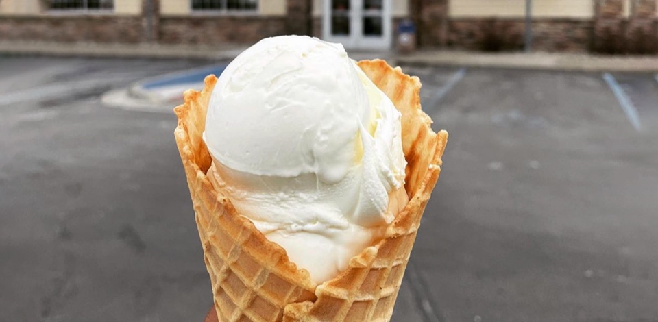 A cone of Vanilla Fresh Frozen Custard is held in front of a Culver's Restaurant.