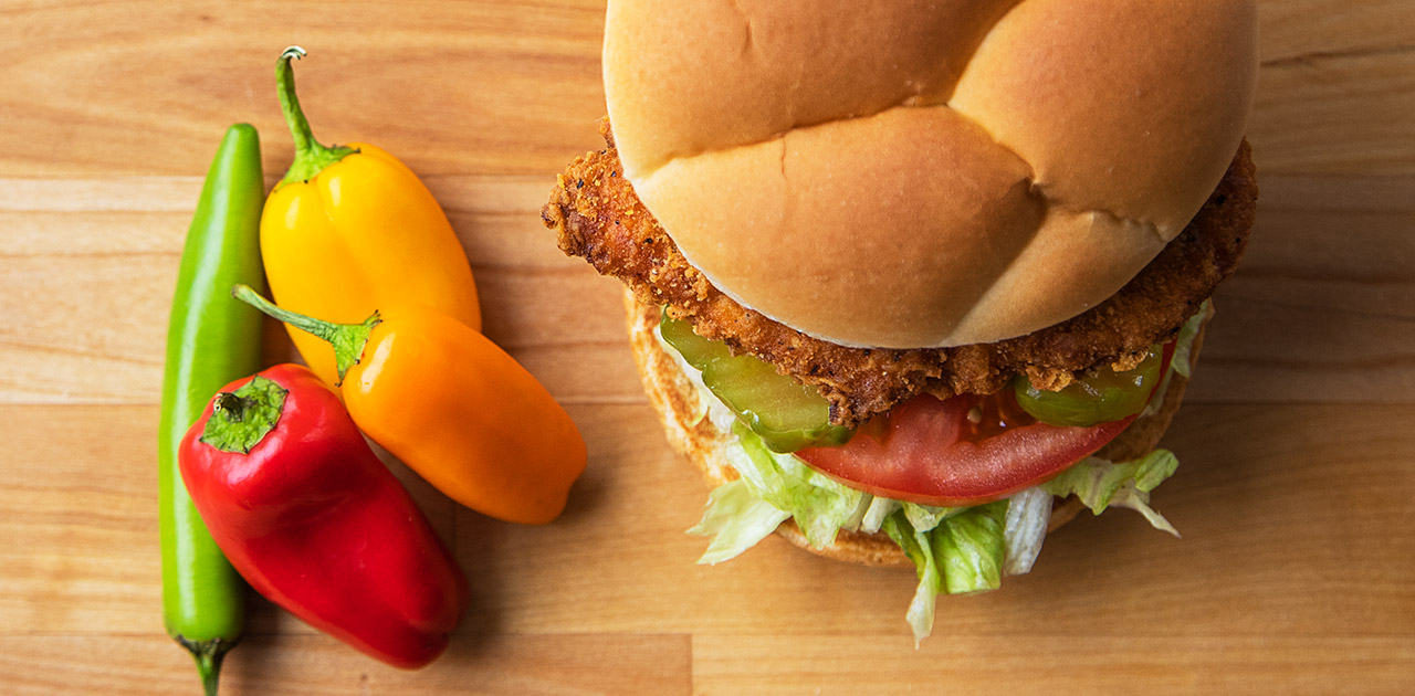 The Spicy Crispy Chicken Sandwich sits next to four peppers on a wood table.