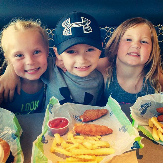 Three toddlers, one boy and two girls, sit in a Culver’s booth with chicken tenders, ketchup and crinkle cut fries in front of them as they smile at the camera