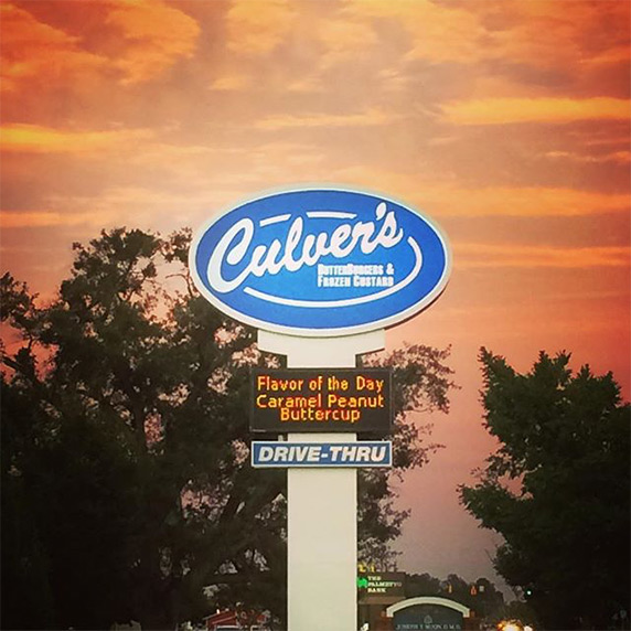 A Culver’s drive-thru sign displaying “Flavor of the Day Caramel Peanut Buttercup” with an orange sunset in the background
