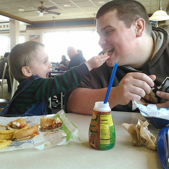 A toddler in overalls laughing while feeding a crinkle cut french fry from Culver’s to a relative sitting next to him