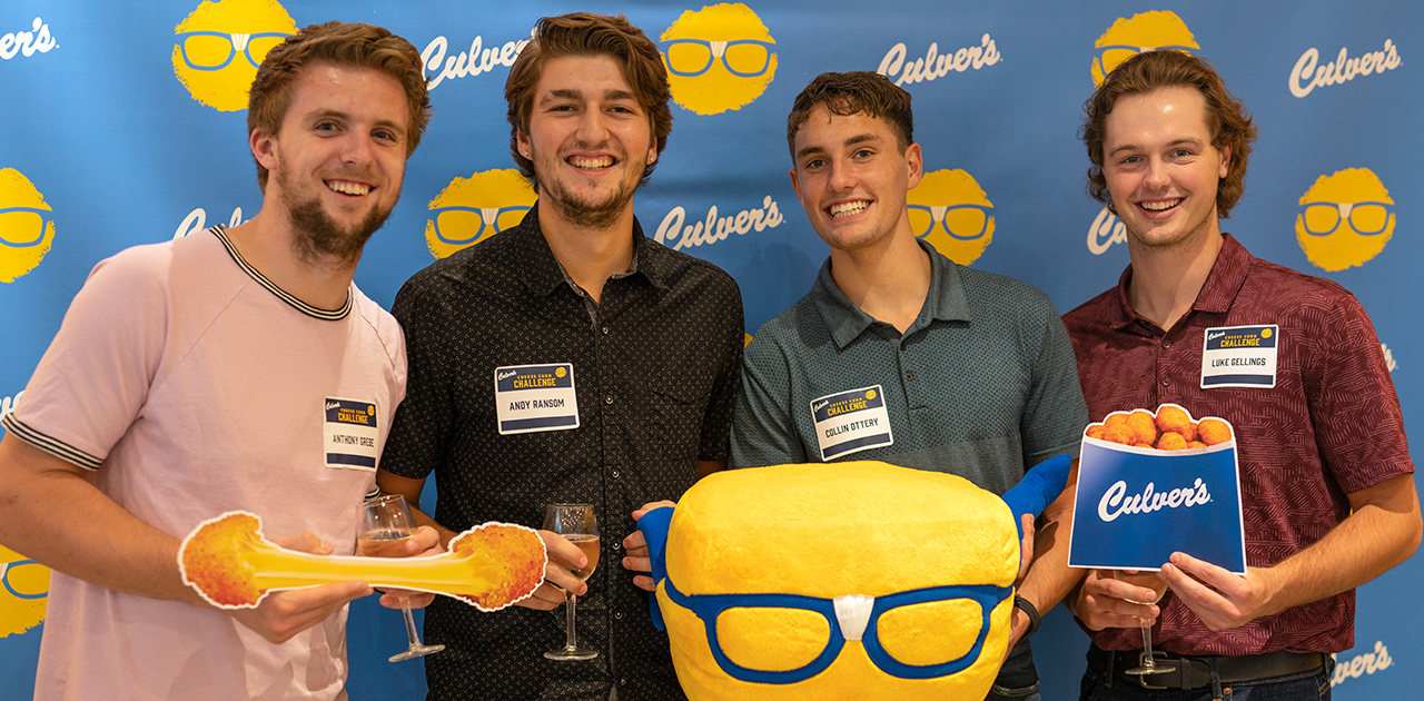 A group of young men take a photo with cheese curd props in front of a Culver’s backdrop