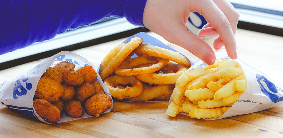 A hand reaches for Onion Rings, Crinkle Cut Fries and Cheese Curds on a table.
