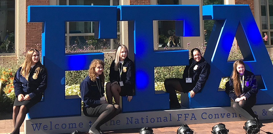 Five FFA Members in blue jackets pose along with a giant FFA sign