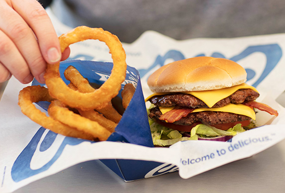 Culver’s Bacon Deluxe shown with Onion Rings in a disposable basket lined with Culver’s paper.