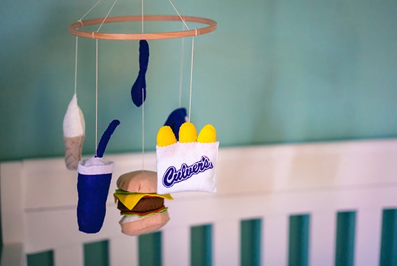 A baby mobile with cheese curds, a drink cup and a ButterBurger hangs over a crib.