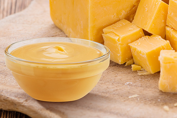 A glass cup of cheese sauce sits on a wood board next to cubes of cheddar cheese