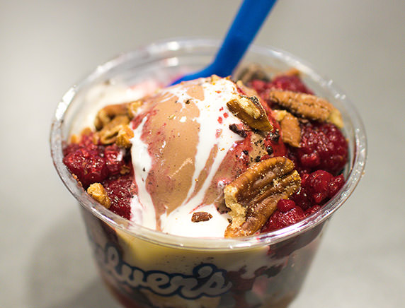 A Sundae with Pecans, Raspberry, Oreo and Marshmallow sits on a table with a blue spoon in the custard.
