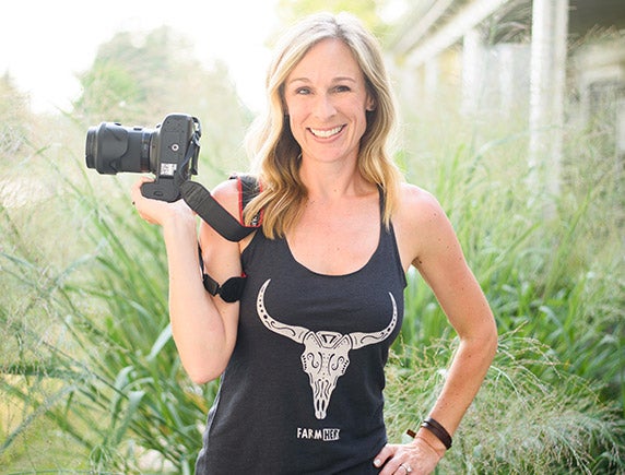 Marji Alaniz stands in a yard with tall grass while holding a camera.