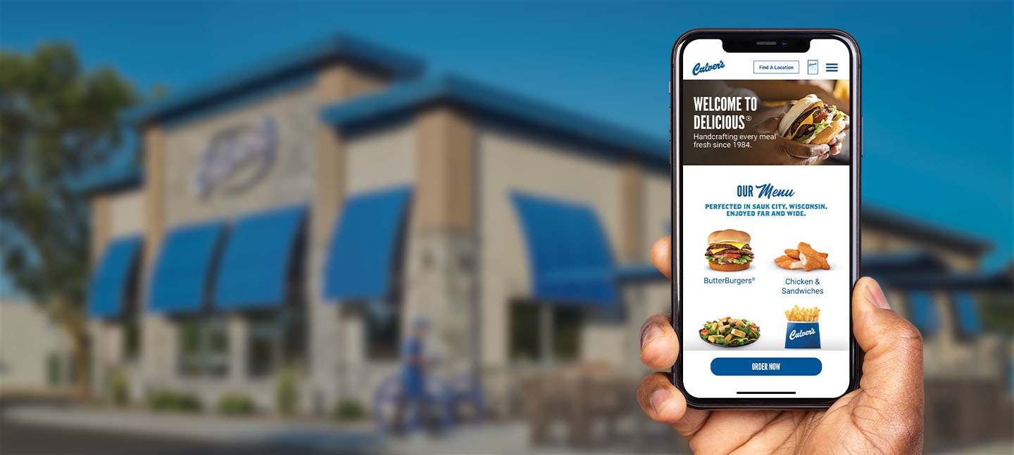 Hand holding an iPhone in the foreground with a blurred Culver's restaurant in the background.