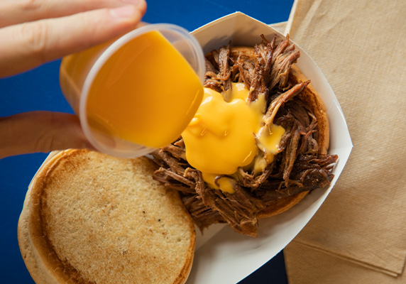 person drizzles cheese sauce on top of beef pot roast sandwich]