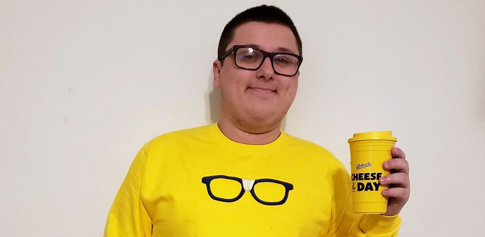 Kurtis Culver wearing a yellow Curd Nerd sweatshirt with black framed glasses, and holding a “Cheese the Day” yellow travel mug