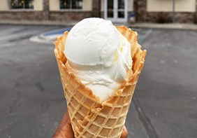 A cone of Vanilla Fresh Frozen Custard is held in front of a Culver's Restaurant.