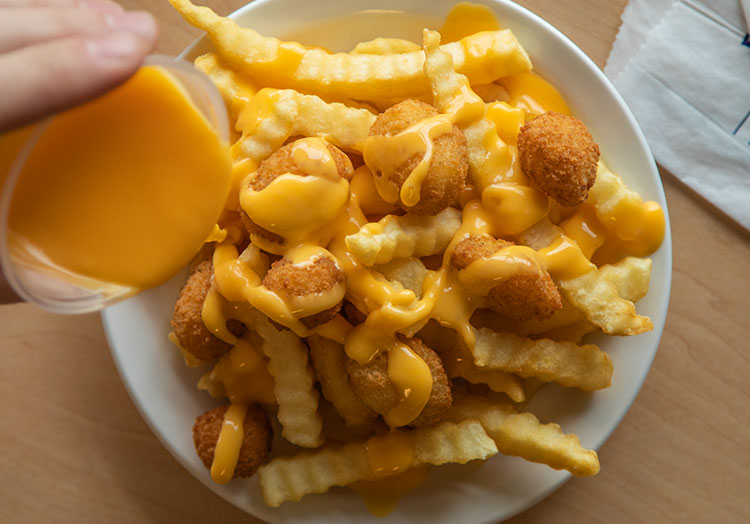 person drizzles cheese sauce over a container of crinkle cut fries and cheese curds