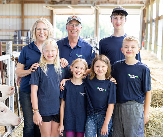 Family smiling and standing in barn next to cow.